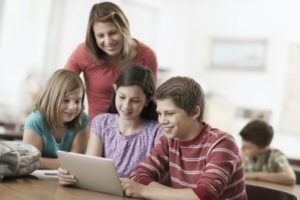 A group of students in class and an adult teacher looking at a digital tablet.