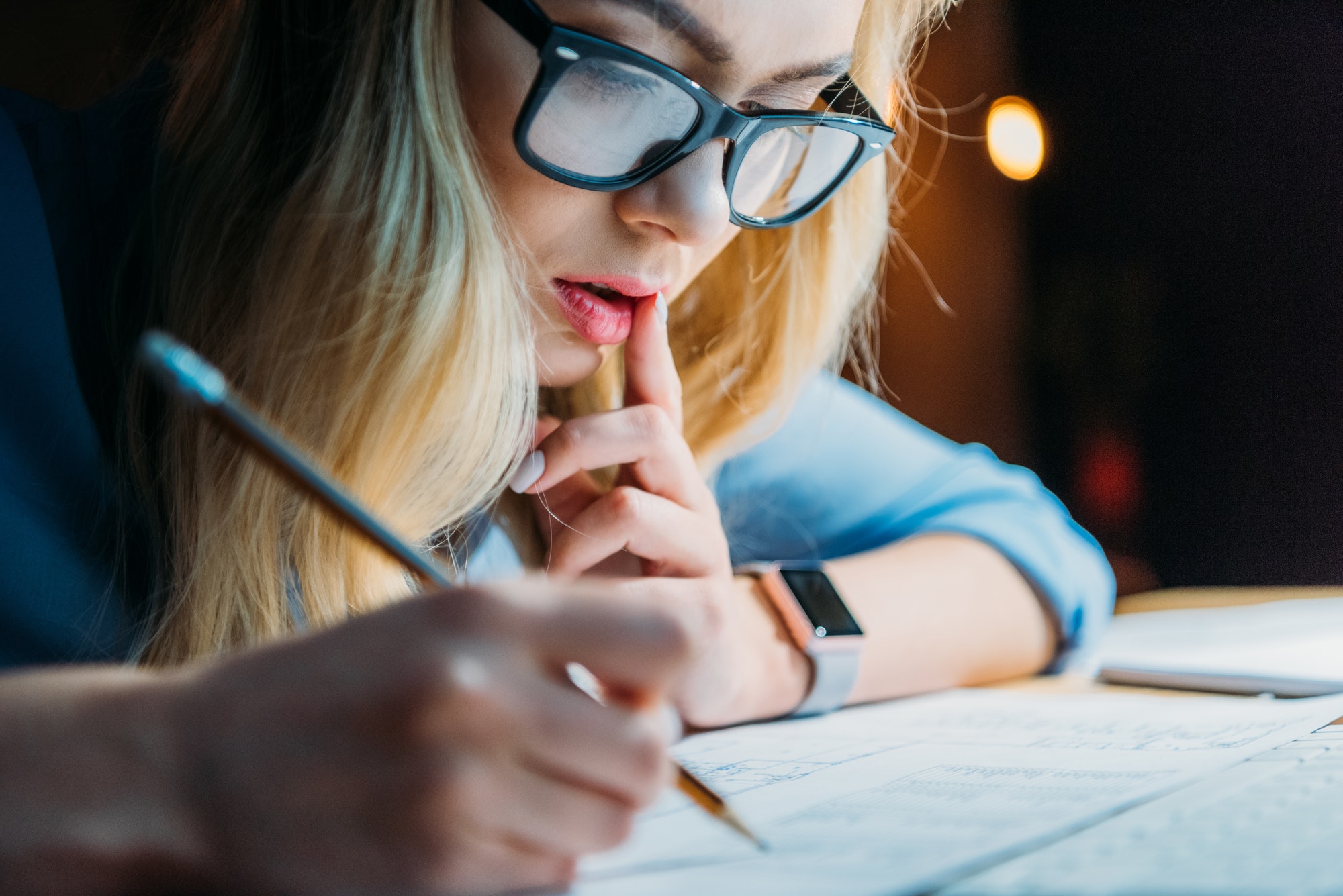 young blonde caucasian student in eyeglasses thinking and writing something with pencil, studying